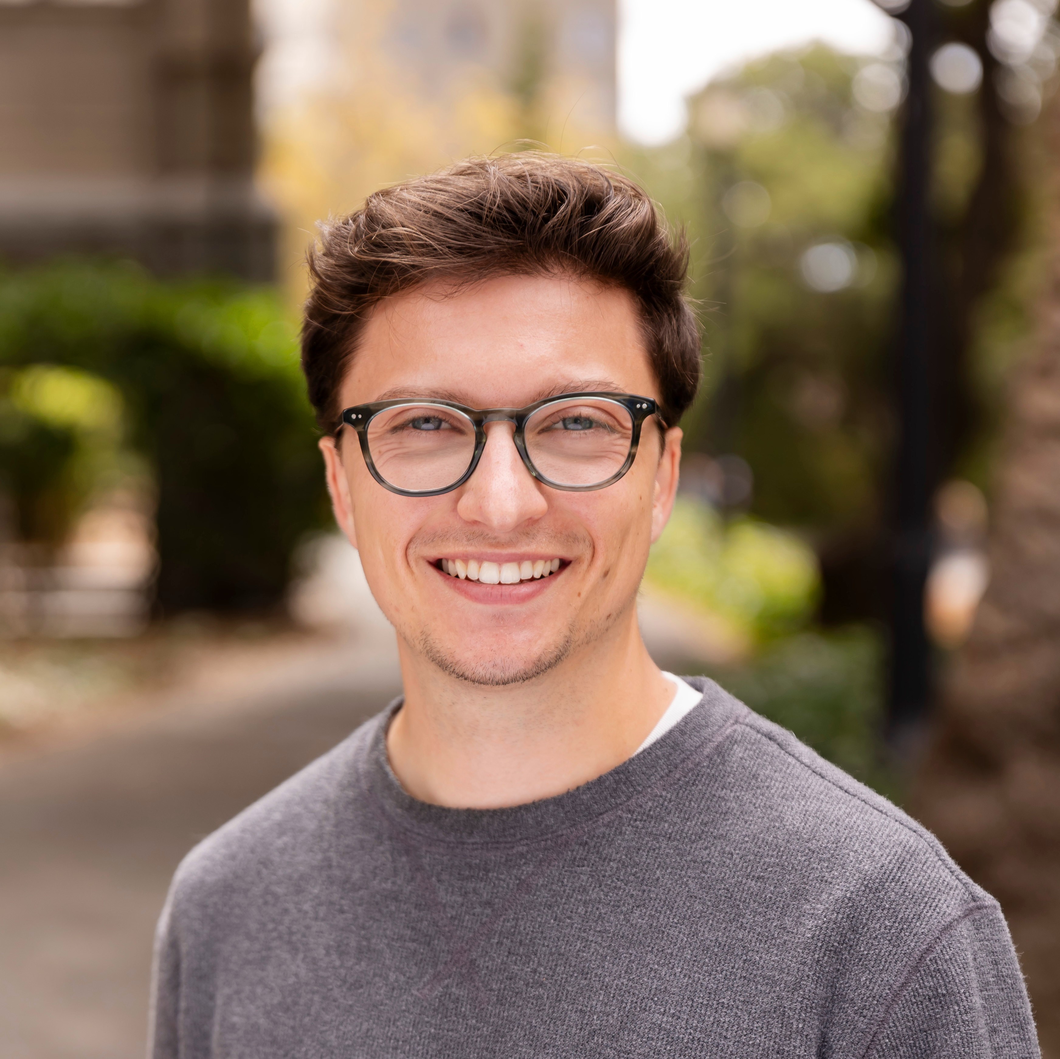 A picture of Chase Stokes. He is wearing a gray sweater, dark green plastic-rimmed glasses, and is grinning at the camera. Behind him is a blurred greenery.